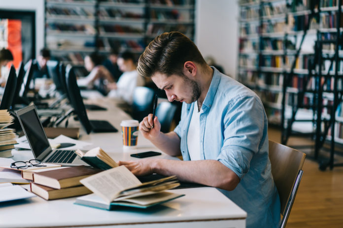 Smart hipster guy sitting in university library doing homework task making research in literature,clever young male student learning for college examination analyzing information from textbooks